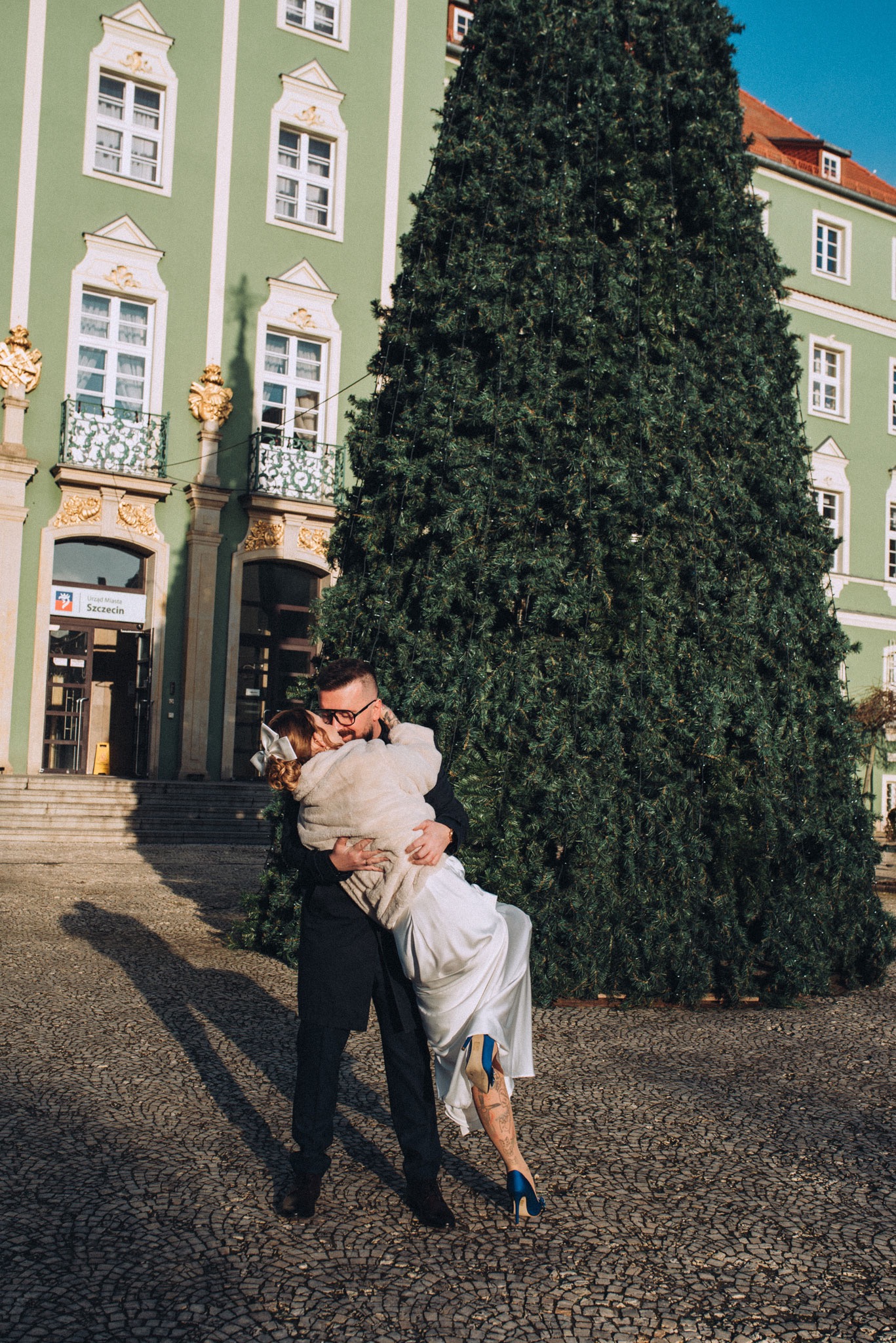 Para młoda w Szczecinie, mężczyzna trzyma pannę młodą na rękach na tle budynku Urzędu Miasta i dużej choinki, panna młoda ma niebieskie buty.