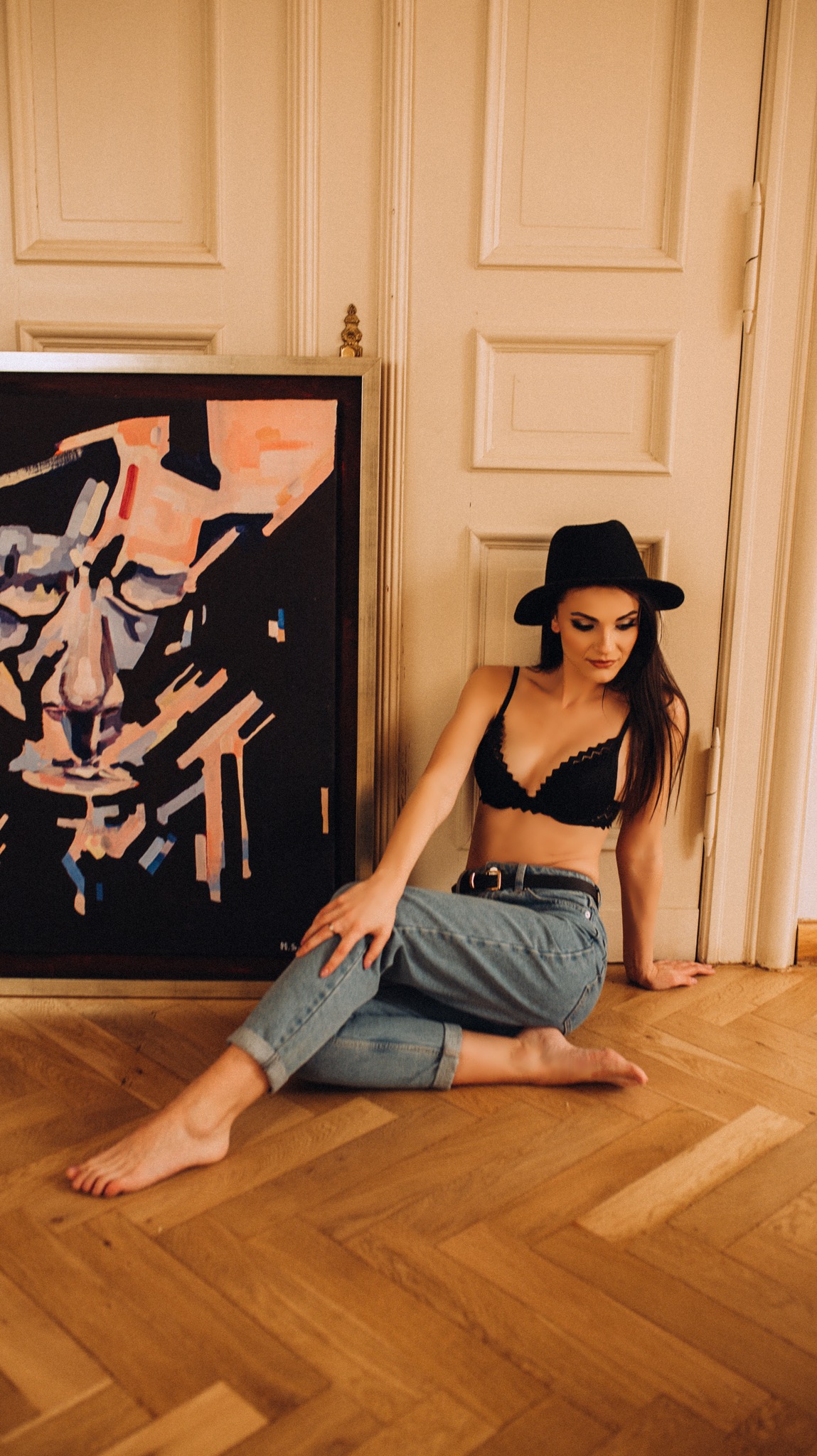 Kobieta w czarnym kapeluszu i koronkowej bieliźnie, siedząca na drewnianej podłodze obok abstrakcyjnego obrazu.