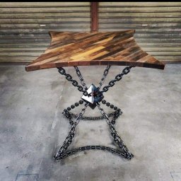 SpawLeks producent meble w stylu Loft usługi spawalnicze metoda TIG spawania aluminium oraz stali - Stół z blatem z desek ułożonych w jodełkę i podstawą z łańcuchów spawanych do metalowej piramidki, na tle metalowej rolety.