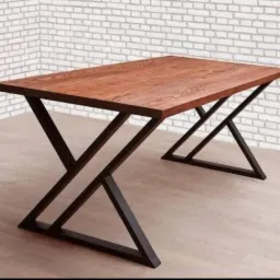 Stół z blatem z drewna o ciepłym odcieniu i metalowymi nogami w kształcie litery X, ustawiony na jasnej podłodze na tle ściany z białej cegły.