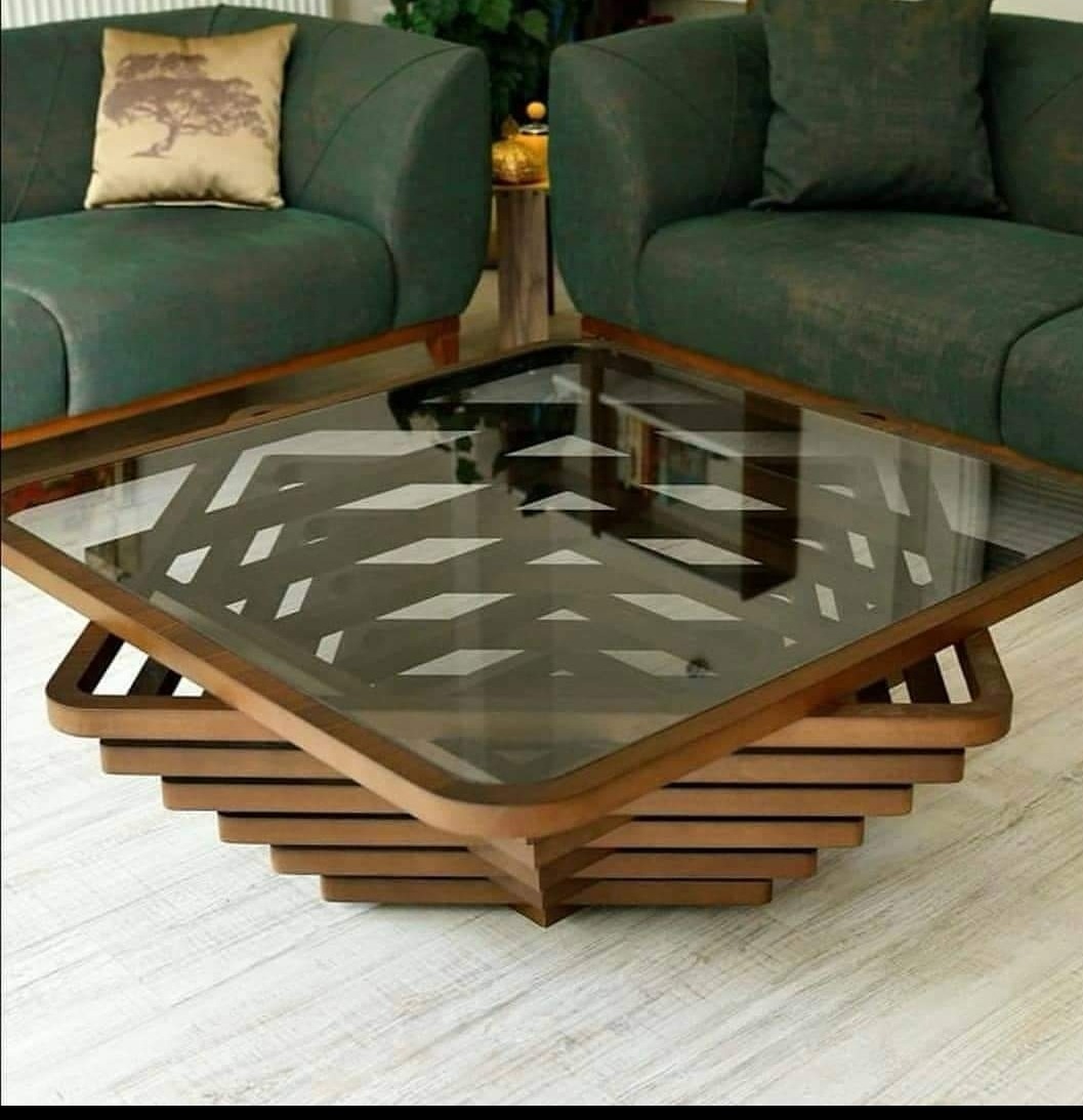 Szklany stolik kawowy z drewnianą podstawą o kaskadowej strukturze, ustawiony w salonie z zielonymi sofami i jasną podłogą.