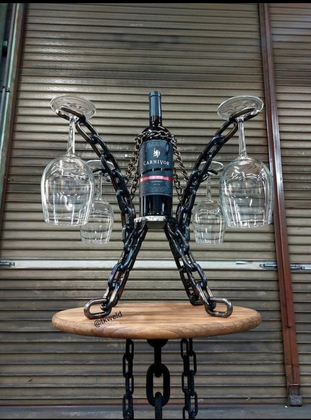 Stojak na wino i kieliszki wykonany z łańcucha spawalniczego, na drewnianym blacie, z butelką wina Carnivor i czterema kieliszkami, na tle metalowej bramy garażowej.
