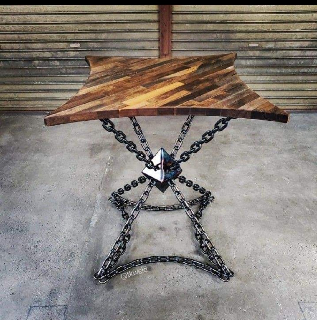 Stół z blatem z desek ułożonych w jodełkę i podstawą z łańcuchów spawanych do metalowej piramidki, na tle metalowej rolety.
