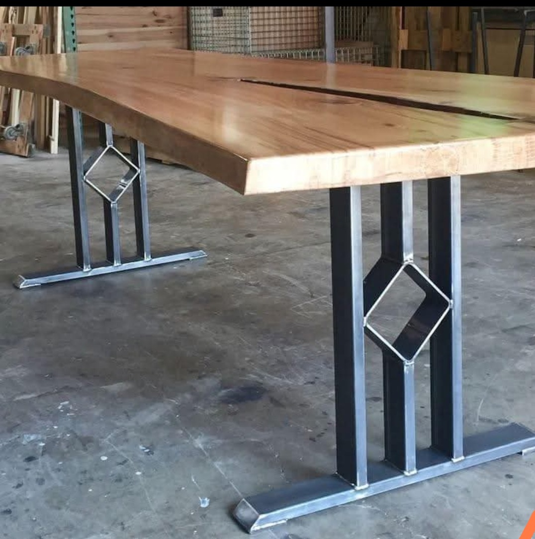 Stół z blatem z jasnego drewna o nieregularnych krawędziach i czarną szczeliną pośrodku, wsparty na metalowych nogach z geometrycznym wzorem rombu.