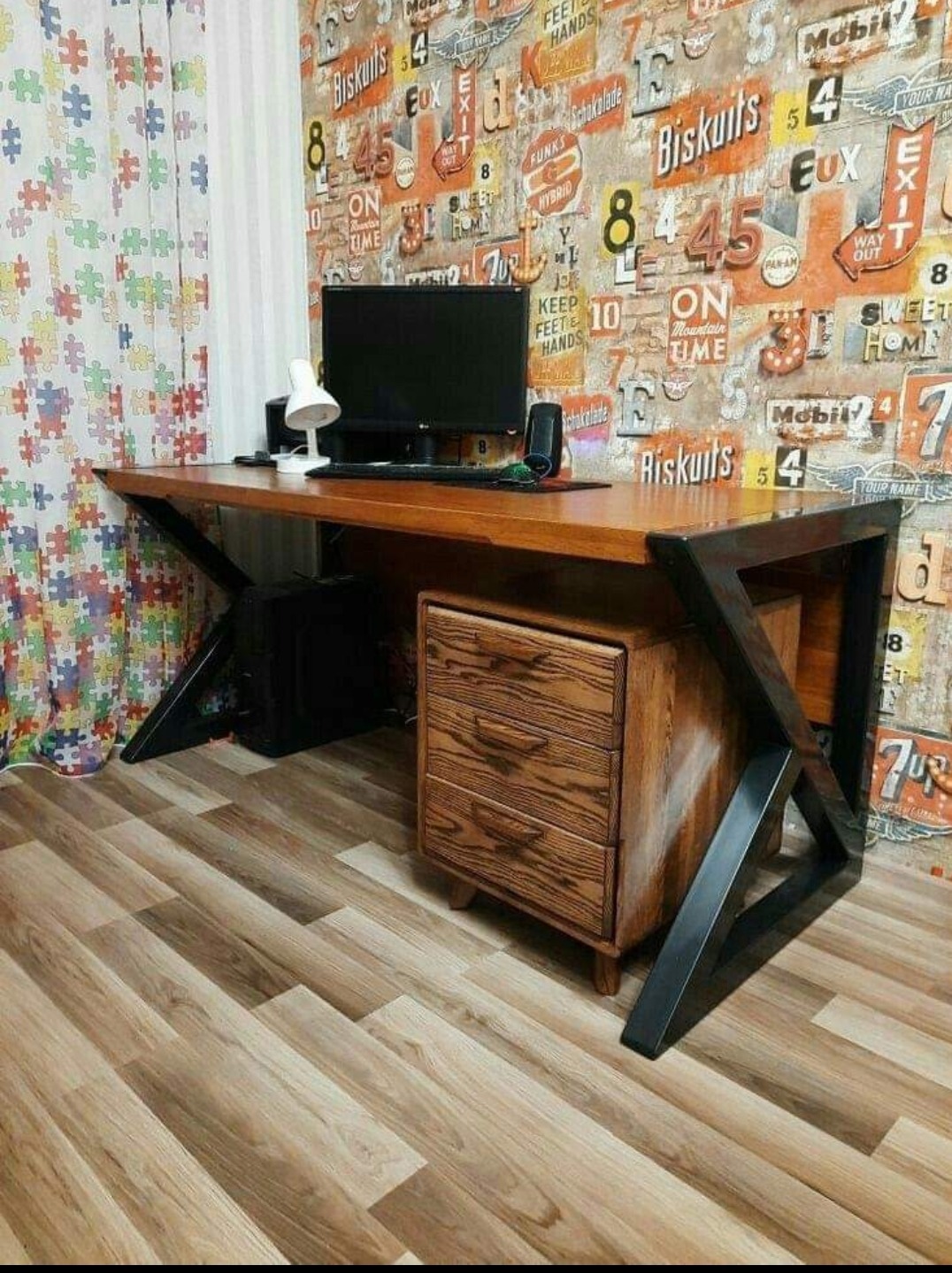 Drewniane biurko z szufladami i metalowymi nogami w stylu industrialnym, na blacie monitor, lampka i głośniki, w tle wzorzysta tapeta i zasłona z motywem puzzli.