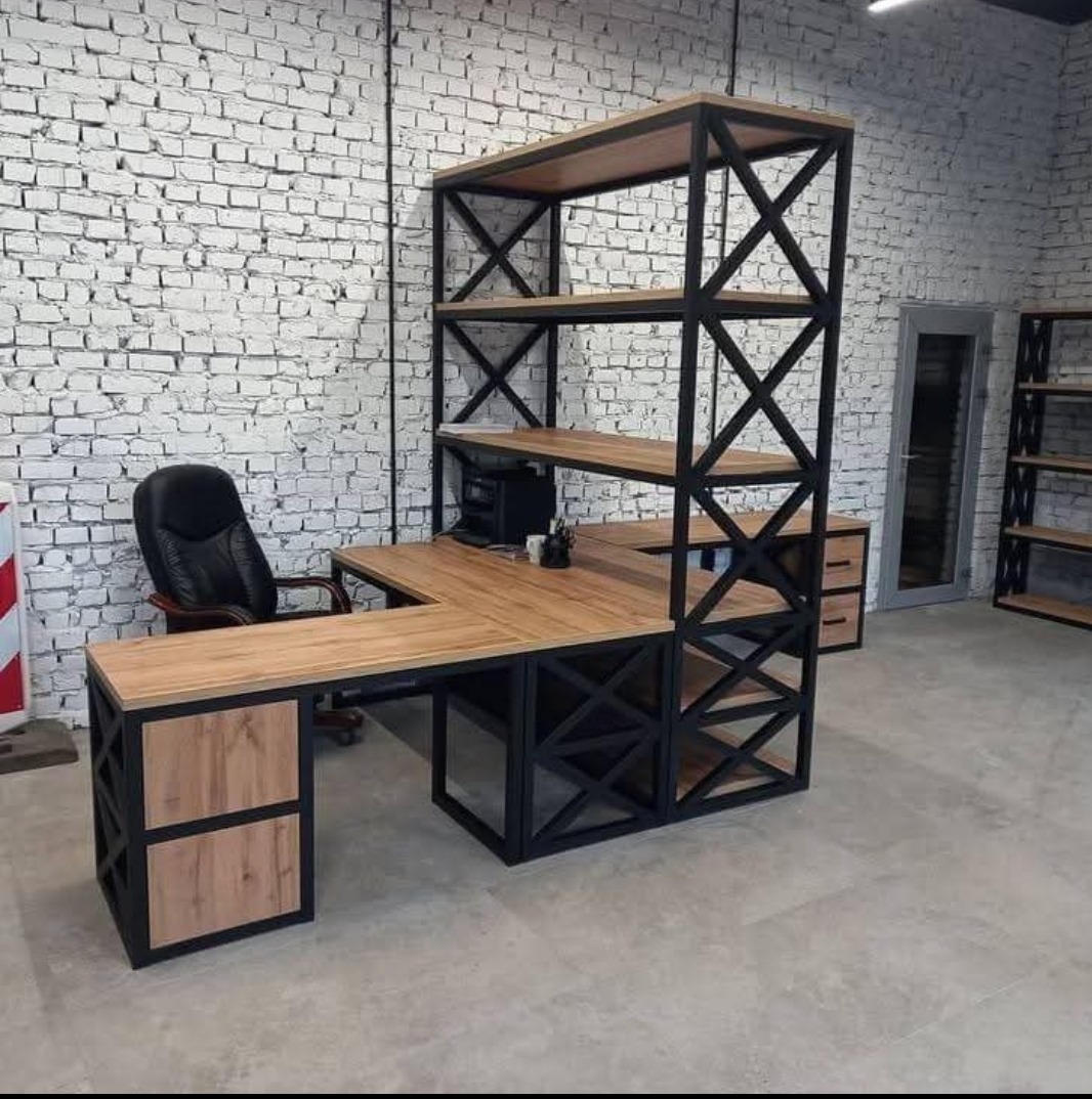 Kątowe biurko z drewnianym blatem i regałem z czarną metalową ramą na tle ściany z białej cegły, widoczne krzesło biurowe i dodatkowy regał w tle.