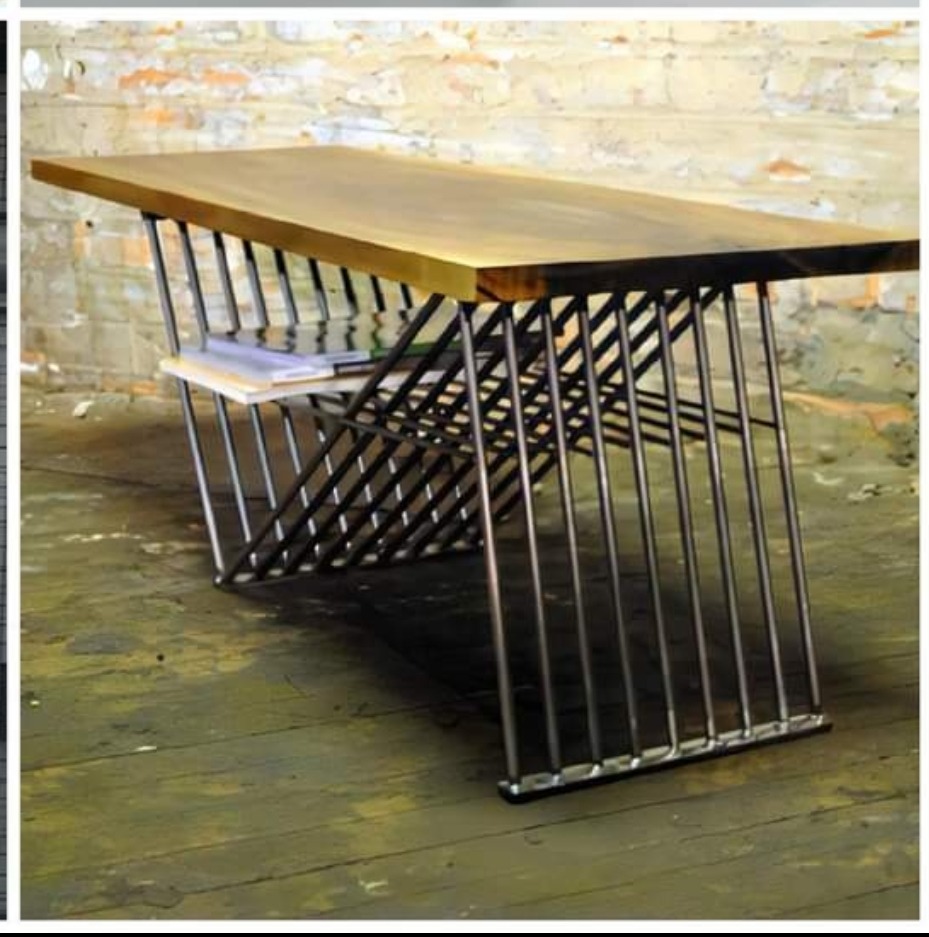 Biurko z blatem z jasnego drewna i metalową podstawą z pionowych prętów, z półką na książki, na tle ceglanej ściany i drewnianej podłogi.