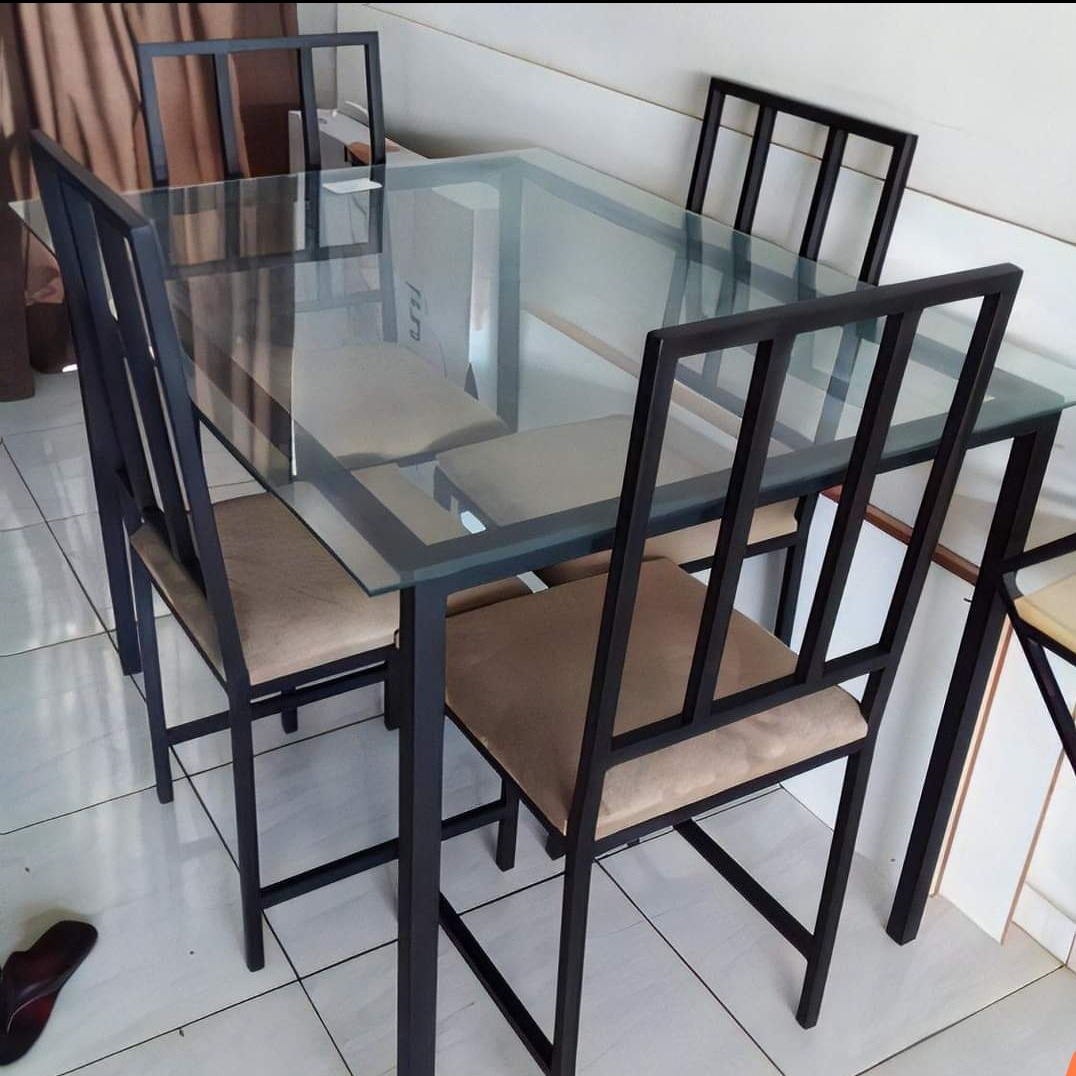 Stół z szklanym blatem i czterema krzesłami z metalowymi ramami i beżowymi siedziskami, widoczne na tle białej ściany i podłogi z jasnych płytek, z jednym butem leżącym na podłodze w lewym dolnym...