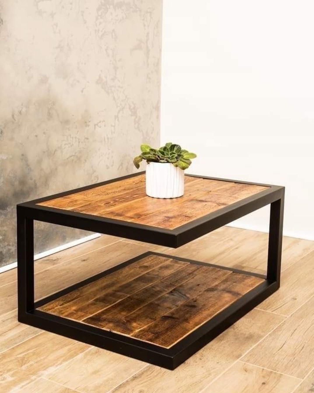 Dwupoziomowy stolik kawowy z drewnianym blatem i czarną metalową ramą, na górnym blacie mała roślina w białej doniczce, tło stanowi ściana z efektem betonu
