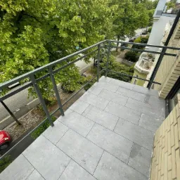 Balkon z szarą, prostokątną terakotą i metalową balustradą w kolorze grafitowym, widok na ulicę z zielenią drzew i fragmentem czerwonego samochodu.