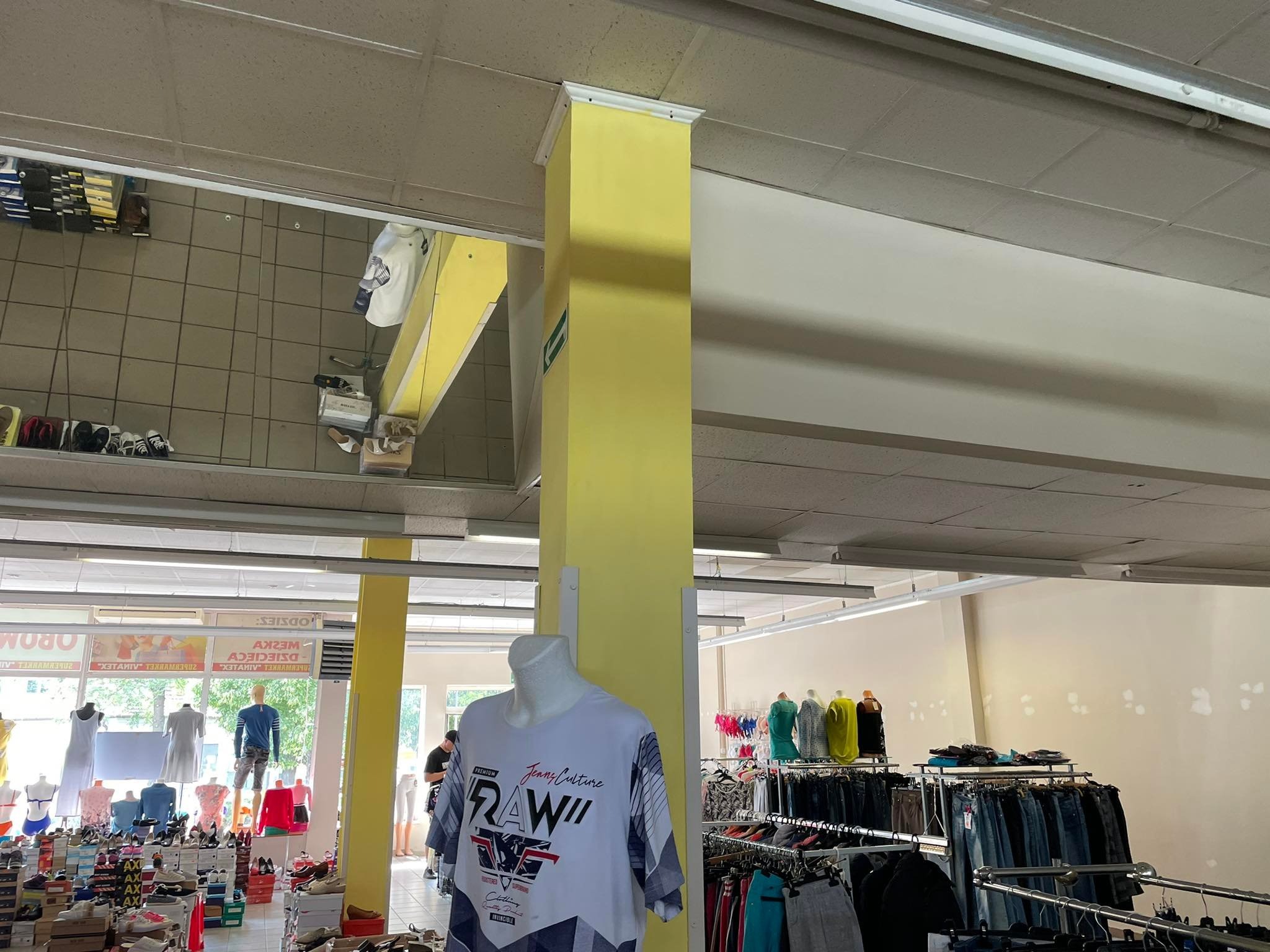 Wnętrze sklepu odzieżowego z widocznymi manekinami, ubraniami na wieszakach, pudełkami z butami i żółtym filarem. Na ścianie widoczne ślady po naprawach.