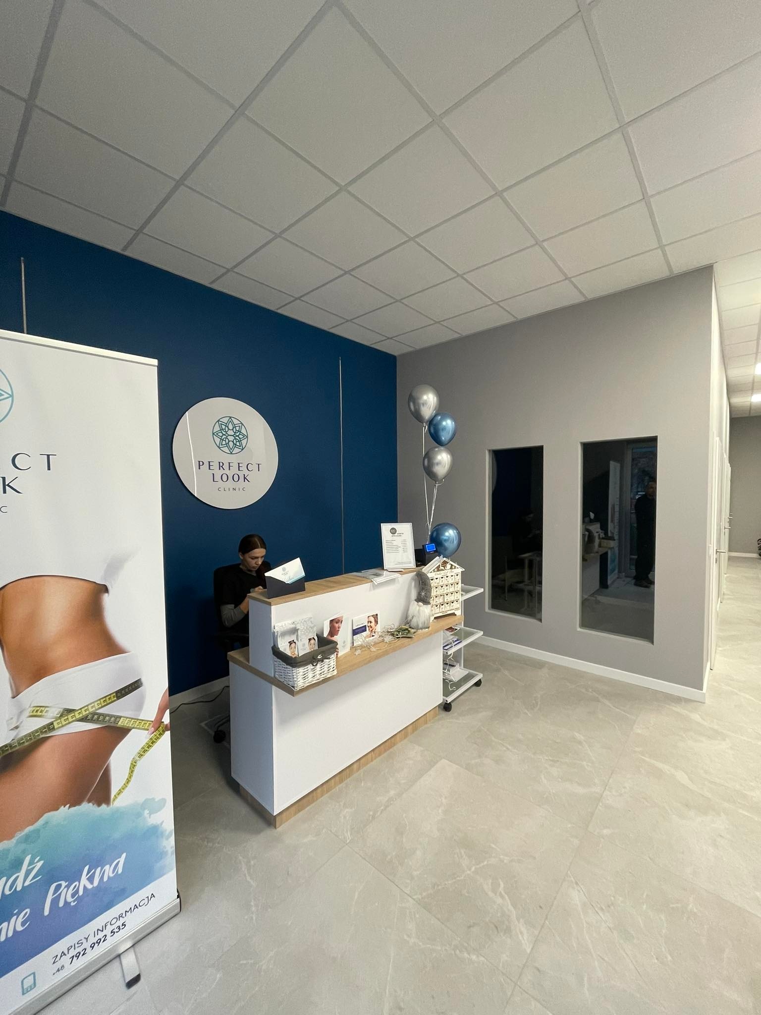 Wnętrze recepcji kliniki medycyny estetycznej z ladą recepcyjną, plakatem reklamowym i dekoracjami, utrzymane w tonacji bieli, szarości i granatu.