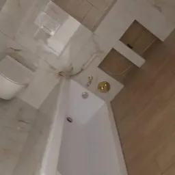 Nowoczesna biała wanna z złotą armaturą w łazience z beżowymi płytkami imitującymi marmur i drewno, widoczne przewody instalacyjne przed wykończeniem.