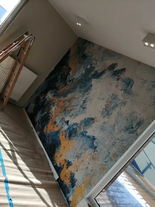 Ściana w pokoju po dekoracyjnym malowaniu, z abstrakcyjnym wzorem w odcieniach błękitu, szarości i złota, drabina malarska w rogu pomieszczenia.