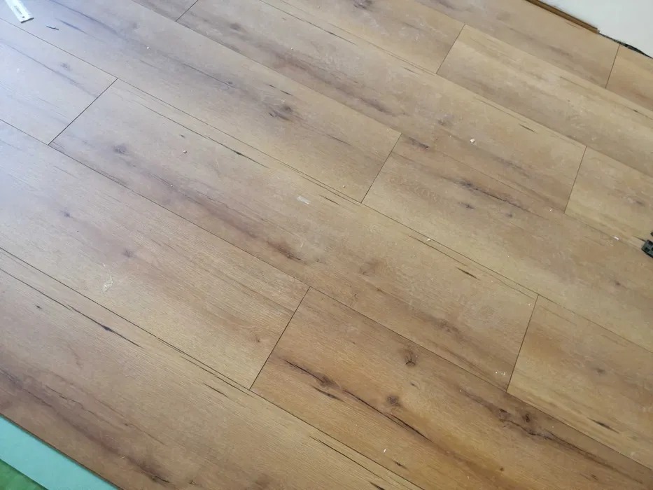 Ułożone panele podłogowe w odcieniach brązu, widoczne drobne zabrudzenia i fragment listwy przypodłogowej w tle.