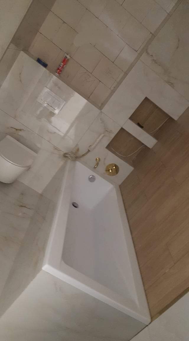 Nowoczesna biała wanna z złotą armaturą w łazience z beżowymi płytkami imitującymi marmur i drewno, widoczne przewody instalacyjne przed wykończeniem.