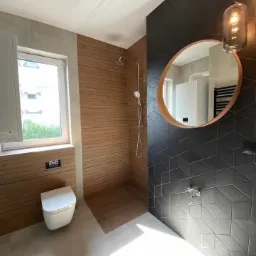 Nowoczesna łazienka z drewnianą okładziną prysznica, czarnymi trójwymiarowymi płytkami, owalnym lustrem i wiszącą lampą z żarówką Edisona.