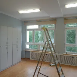 Pok&oacute;j z jasnymi szafkami i świeżo pomalowanymi ścianami na szaro-niebieski kolor, z rozstawioną aluminiową drabiną na pierwszym planie, okna z roletami i widokiem na zieleń na zewnątrz,...