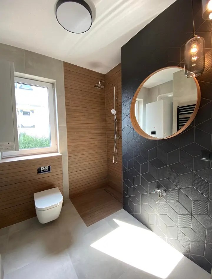 Nowoczesna łazienka z drewnianą okładziną prysznica, czarnymi trójwymiarowymi płytkami, owalnym lustrem i wiszącą lampą z żarówką Edisona.