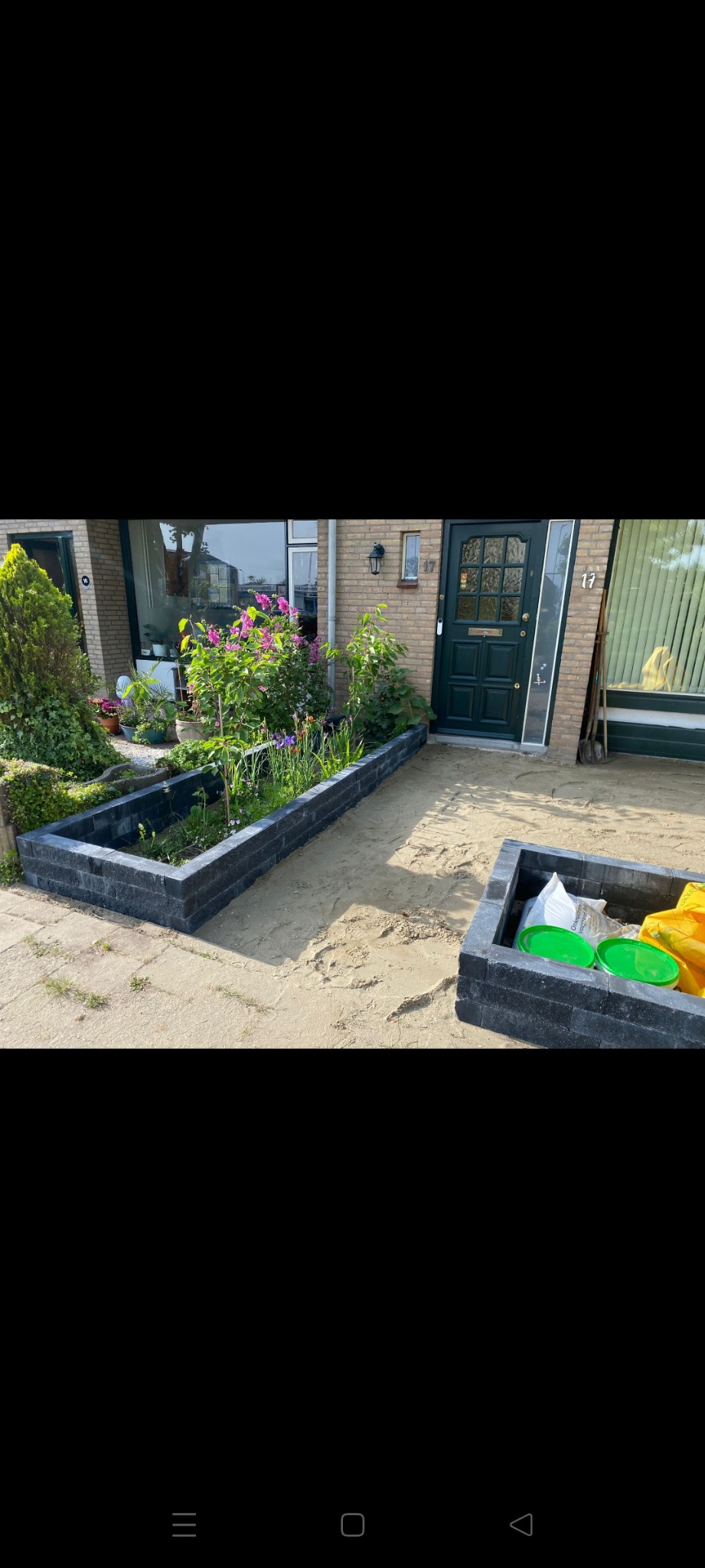 Kwadratowa rabata z ciemnych cegieł, wewnątrz kwiaty i rośliny, obok rabaty z narzędziami ogrodniczymi i workiem ziemi, na piaszczystym podłożu przed domem.