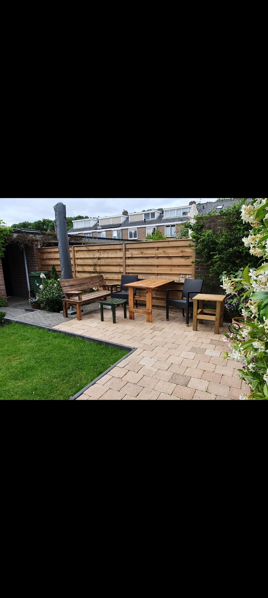 Umeblowany taras z kostki brukowej z drewnianym stołem, ławką, czarnymi krzesłami i zielonym stolikiem, otoczony trawnikiem i drewnianym płotem, kwitnący krzew w prawym dolnym rogu.