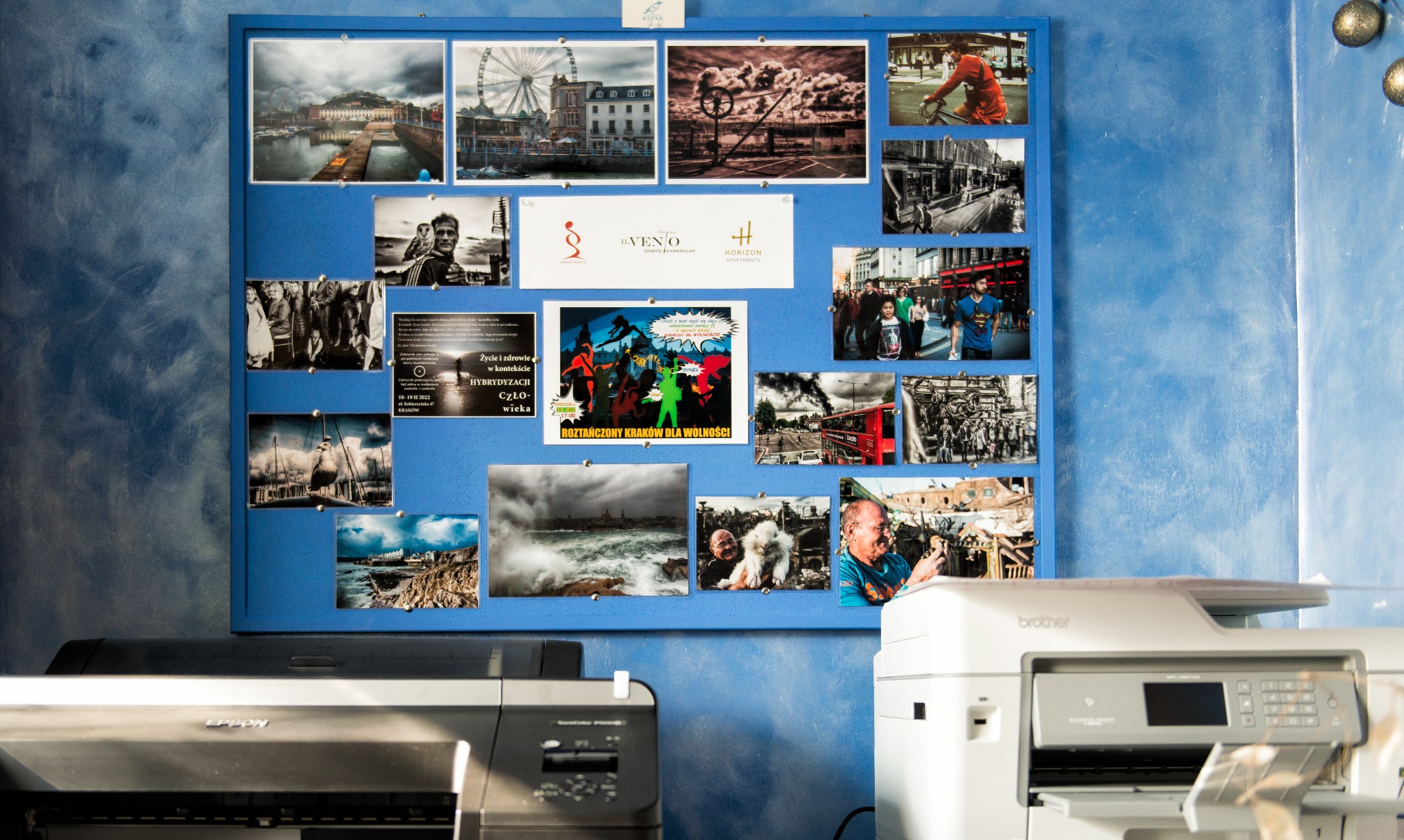 Tablica korkowa z przypiętymi zdjęciami i ulotkami, w tle niebieska ściana z efektem chmur, na pierwszym planie dwie drukarki biurowe.