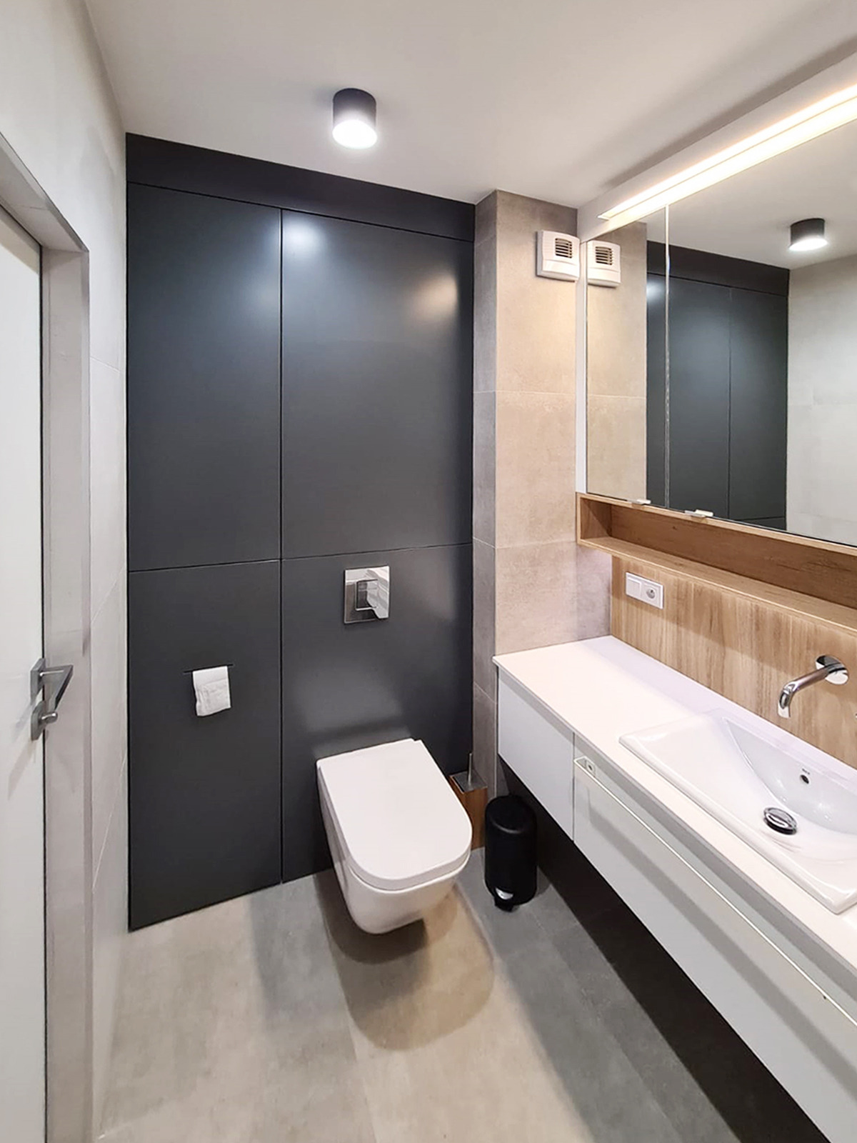 Nowoczesna łazienka z szarą zabudową, podwieszaną toaletą i białą umywalką z szafką, oświetlona podłużnym ledem nad lustrem.