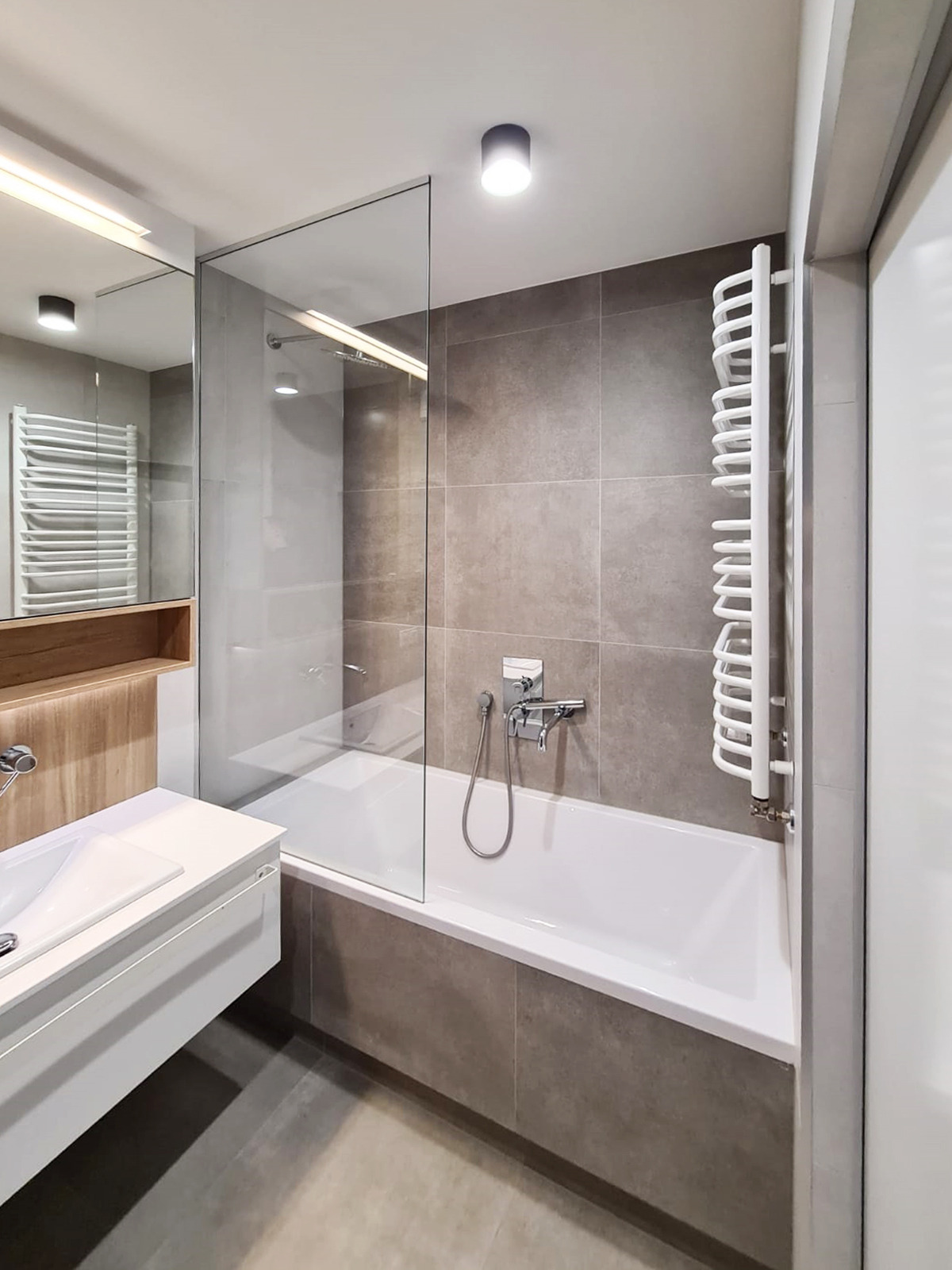 Nowoczesna łazienka z wanną obudowaną szarymi płytkami, szklanym parawanem nawannowym, białym grzejnikiem drabinkowym i umywalką z szafką w minimalistycznym stylu.