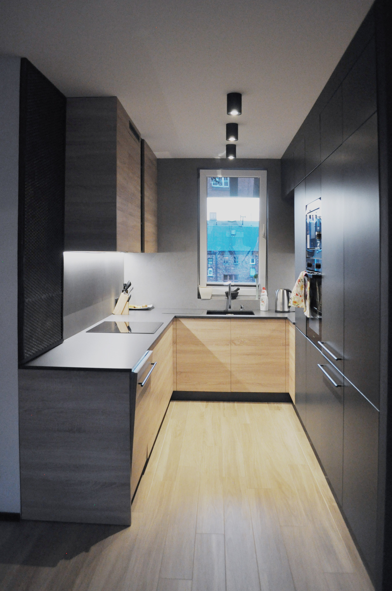 Wąska kuchnia w szarościach i drewnie z oknem na budynek mieszkalny; widoczne czarne, punktowe lampy sufitowe i akcesoria kuchenne na blacie.