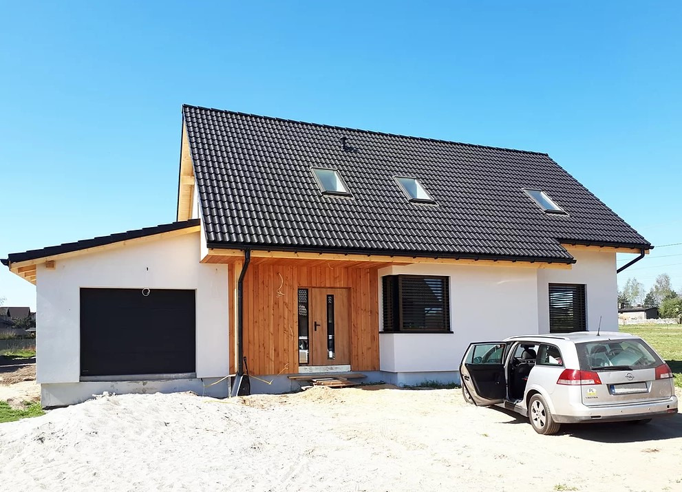 Nowoczesny dom jednorodzinny z czarną dachówką, drewnianą elewacją przy wejściu i otwartymi drzwiami samochodu kombi zaparkowanego obok.