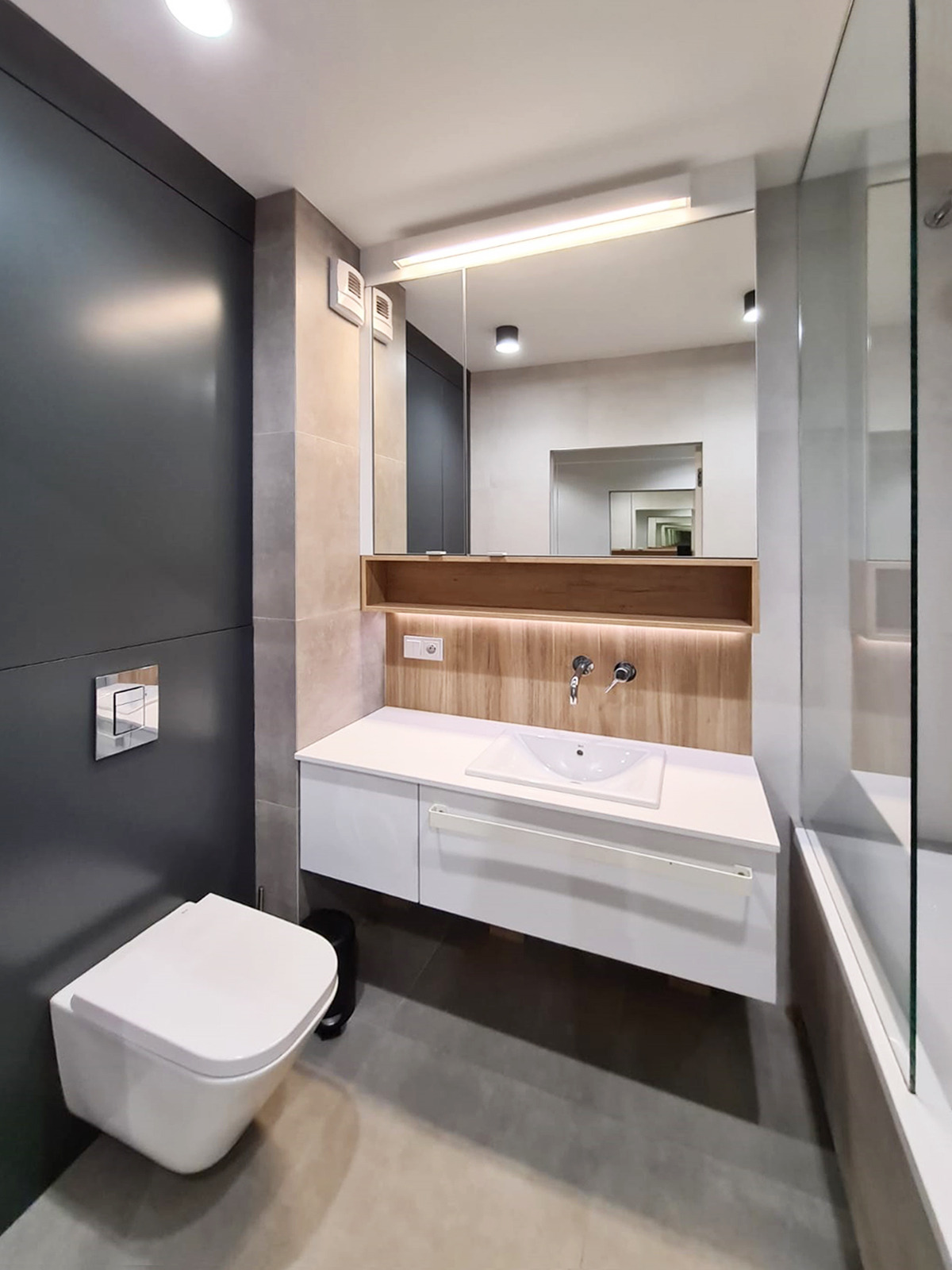 Nowoczesna łazienka z podwieszaną toaletą, białą szafką z umywalką i lustrem z oświetleniem LED, wykończona szarymi i drewnopodobnymi płytkami.