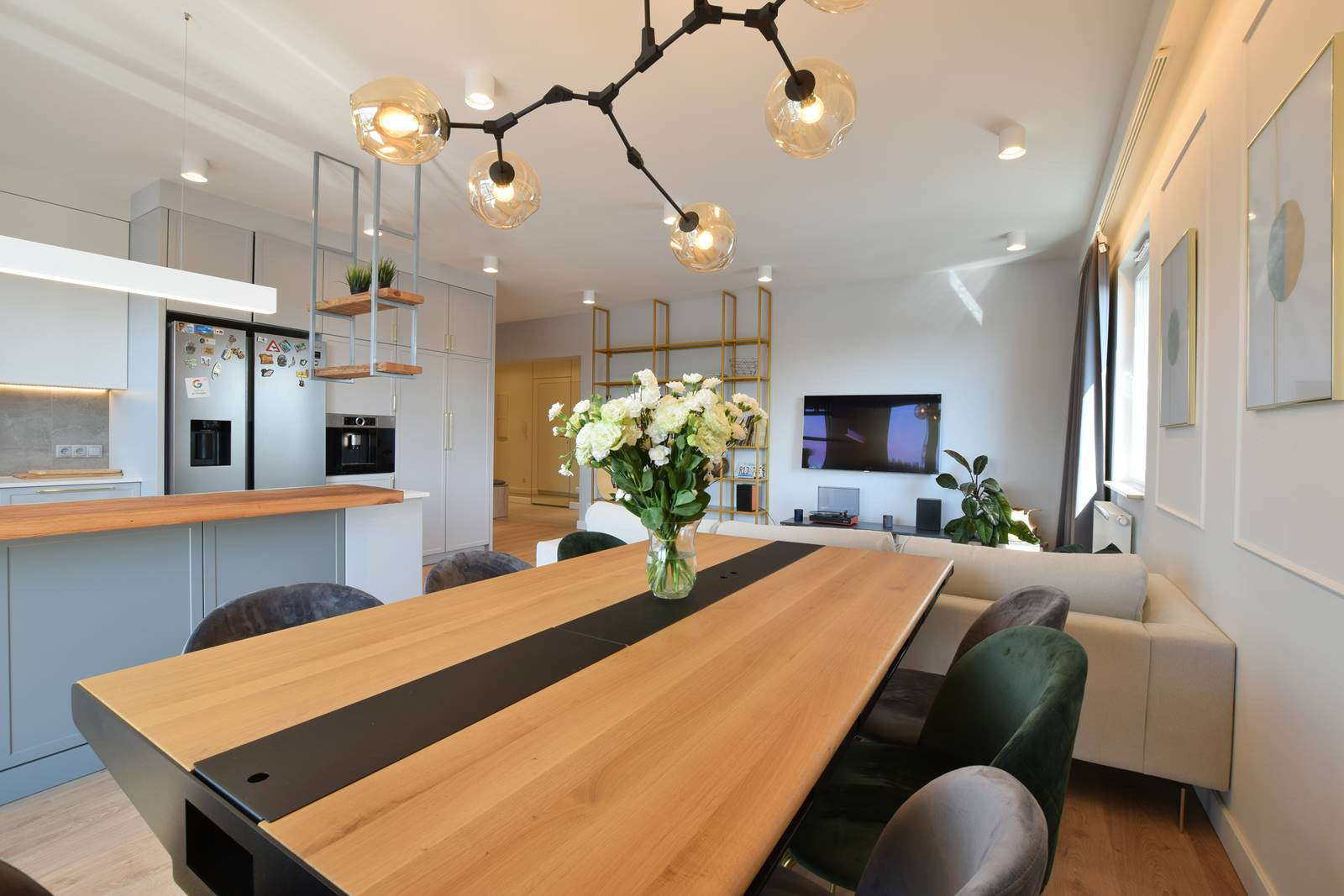 Jasne, przestronne wnętrze: kuchnia z wyspą przechodząca w jadalnię z dużym drewnianym stołem z czarnym pasem pośrodku, nad stołem designerska lampa z kulistymi kloszami, w tle salon z telewizorem...