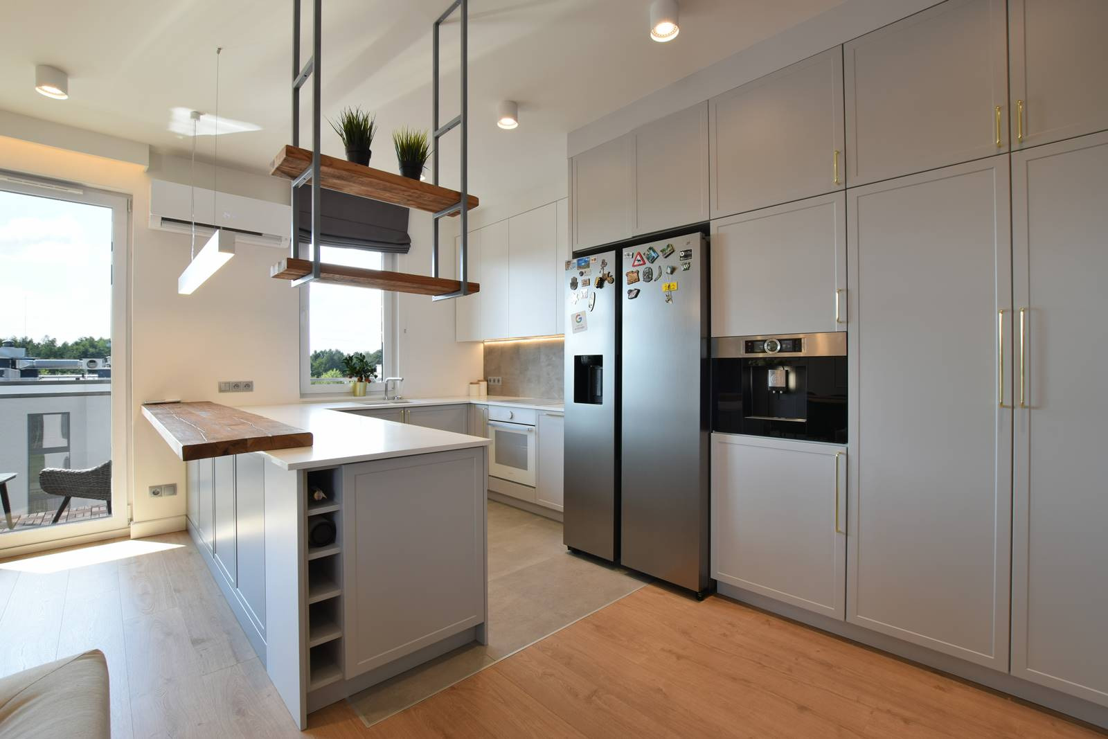 Jasna kuchnia w nowoczesnym stylu z wyspą, drewnianym blatem i półkami wiszącymi pod sufitem, widok na balkon.