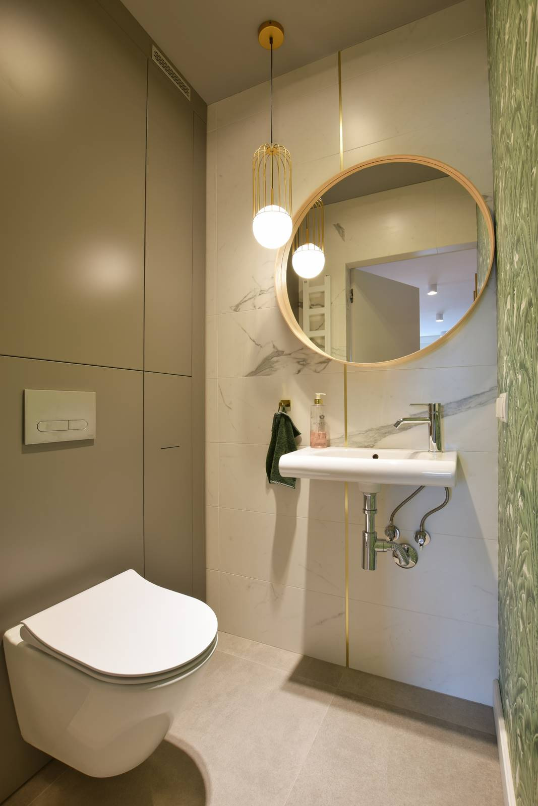 Nowoczesna łazienka z wiszącą toaletą, umywalką i okrągłym lustrem, oświetlona złotą lampą wiszącą, z akcentami marmuru i zielonych wzorów na ścianie.