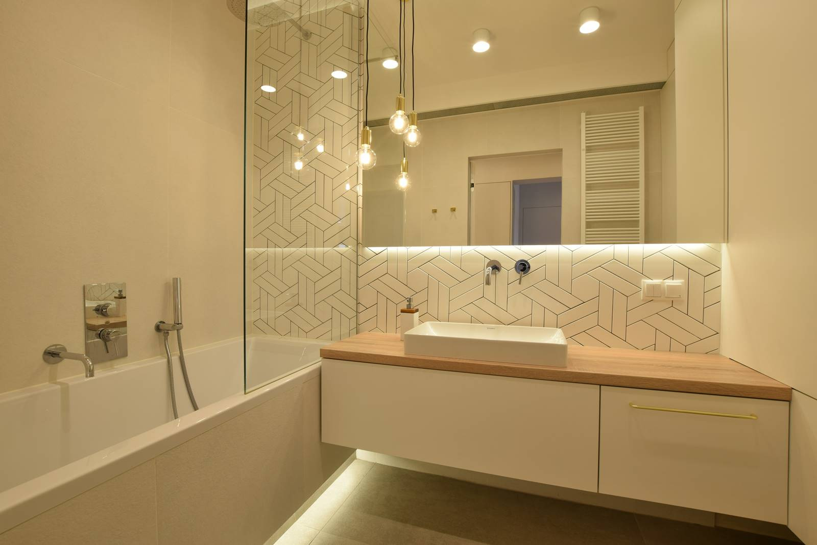 Nowoczesna łazienka z wanną, umywalką nablatową na drewnianym blacie i geometrycznymi płytkami w strefie prysznica, oświetlona wiszącymi lampami z żarówkami Edison.
