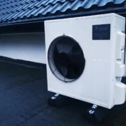 Biała, zewnętrzna jednostka z wentylatorem na czarnym, płaskim dachu z widocznym fragmentem ciemnej dachówki w tle.