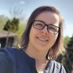 Uśmiechnięta kobieta w okularach na tle rozmytego krajobrazu z domem i zielenią, wykonująca selfie.