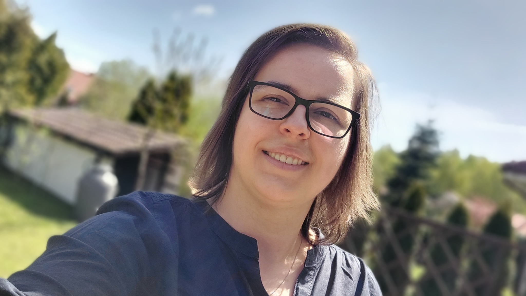 Uśmiechnięta kobieta w okularach na tle rozmytego krajobrazu z domem i zielenią, wykonująca selfie.