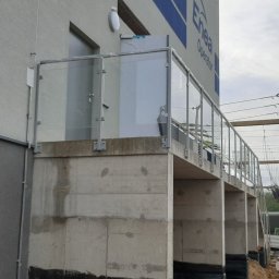 Anatsteel Sp. z o. o. - Zewnętrzne balustrady szklane z metalowymi słupkami, zamontowane na betonowym podeście przy budynku Enea Operator, widoczne elementy konstrukcji i instalacje elektryczne w tle.