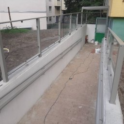 Anatsteel Sp. z o. o. - Zewnętrzna rampa z szarą metalową balustradą i szklanymi panelami, widoczne niedociągnięcia w wykończeniu, w tle budynek mieszkalny i sprzęt budowlany.