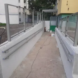 Zewnętrzna rampa dla niepełnosprawnych z szarą, metalową balustradą szklaną i podwójną poręczą, widoczne ślady prac budowlanych, w tle budynek mieszkalny.