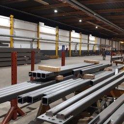 Anatsteel Sp. z o. o. - Stalowe profile kwadratowe, ułożone na podporach w hali produkcyjnej, w tle widoczny wózek widłowy i pracownik.
