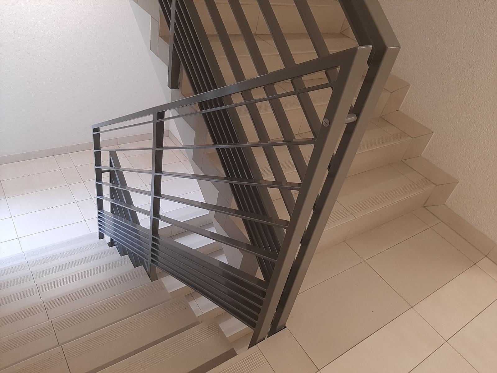 Nowoczesna, metalowa balustrada schodowa z poziomymi prętami, zamontowana na schodach wyłożonych jasnymi płytkami ceramicznymi, widok z góry.