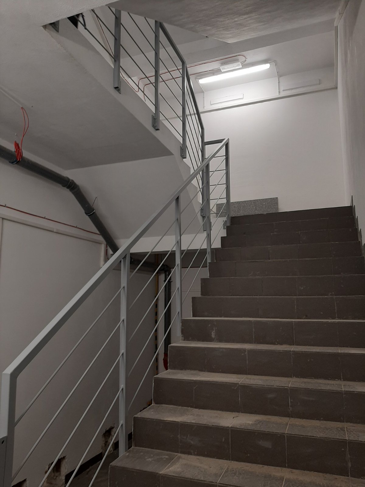 Klatka schodowa z szarymi, płytkowymi schodami i metalową, pomalowaną na szaro balustradą z poziomymi prętami. Widoczne instalacje elektryczne i rury na ścianie.