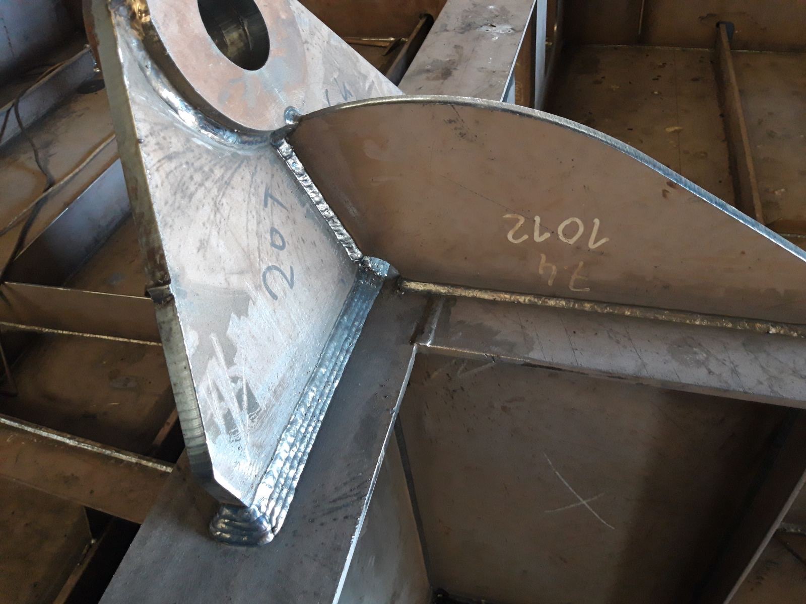 Detal spawanej konstrukcji stalowej z widocznymi śladami po spawaniu i oznaczeniami naniesionymi kredą.