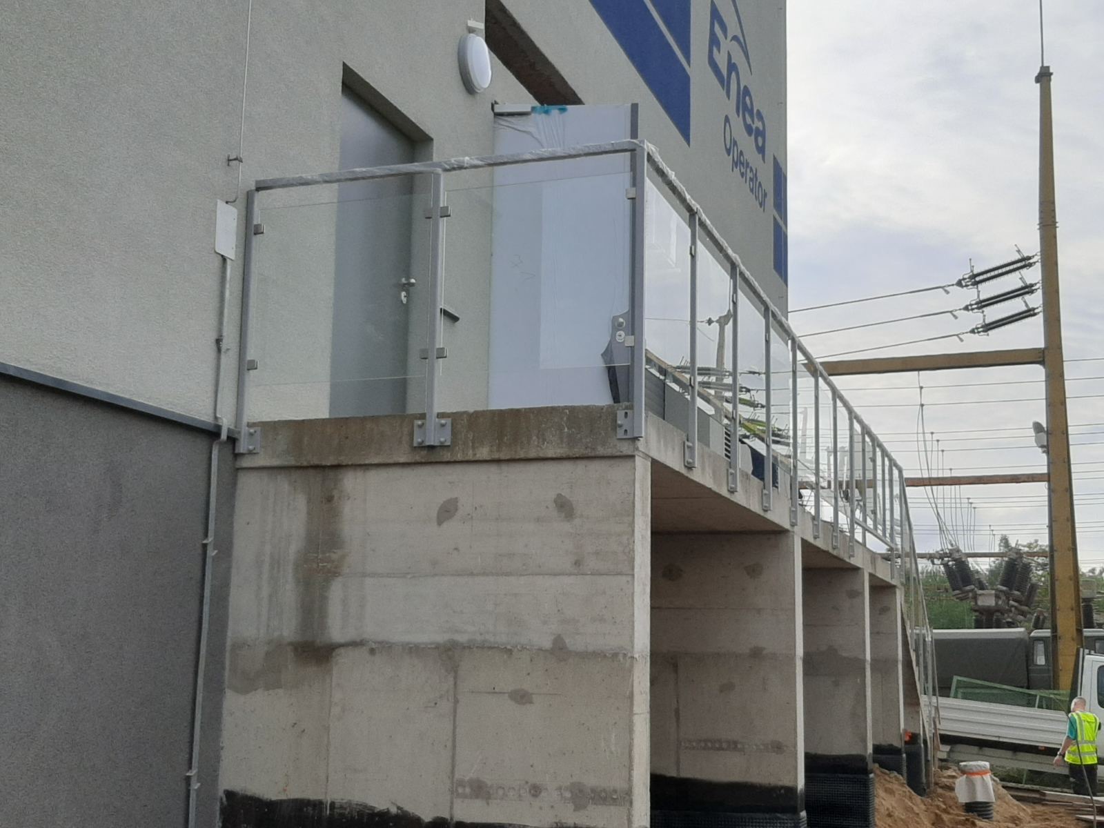 Zewnętrzne balustrady szklane z metalowymi słupkami, zamontowane na betonowym podeście przy budynku Enea Operator, widoczne elementy konstrukcji i instalacje elektryczne w tle.