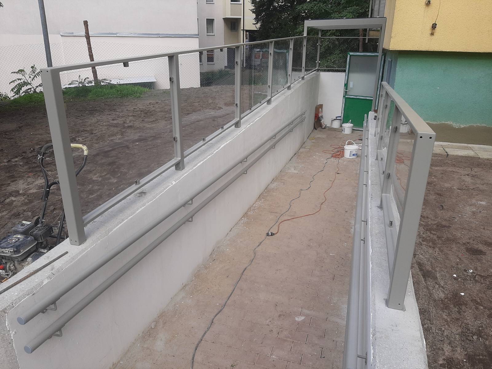 Zewnętrzna rampa z szarą metalową balustradą i szklanymi panelami, widoczne niedociągnięcia w wykończeniu, w tle budynek mieszkalny i sprzęt budowlany.