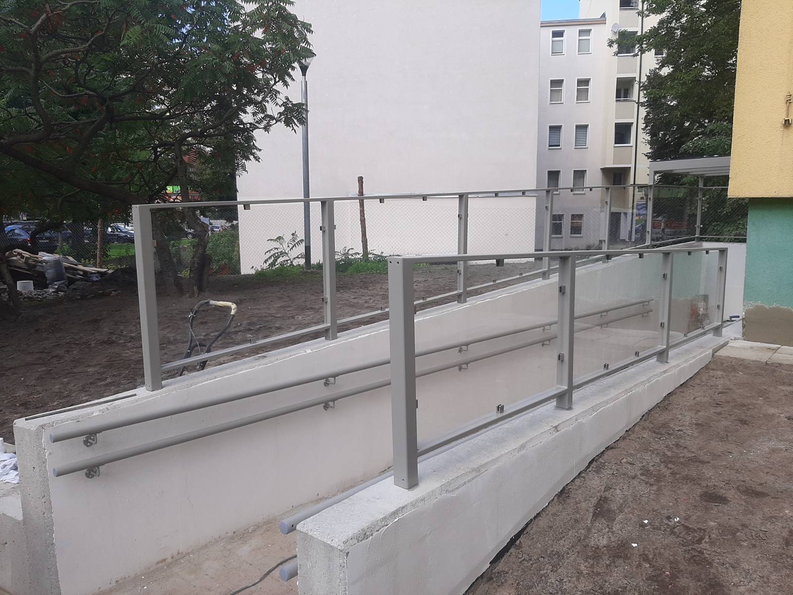 Zewnętrzna rampa dla niepełnosprawnych z szarą metalową balustradą i częściowo szklanym wypełnieniem, widok na budynek mieszkalny w tle.