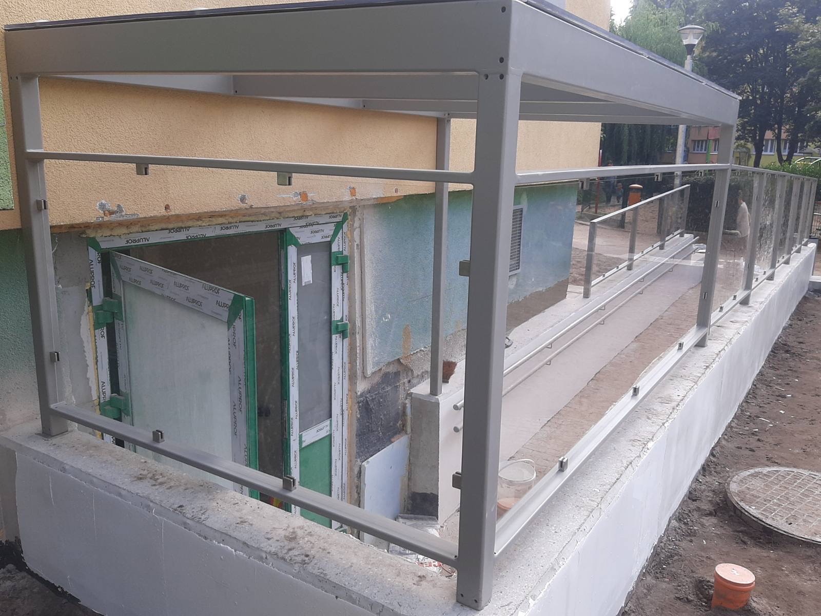 Zewnętrzne schody ze szklanymi balustradami i zadaszeniem, prowadzące do piwnicy budynku mieszkalnego. Widoczne elementy konstrukcyjne z metalu pomalowanego na szaro. Częściowo otwarte drzwi...