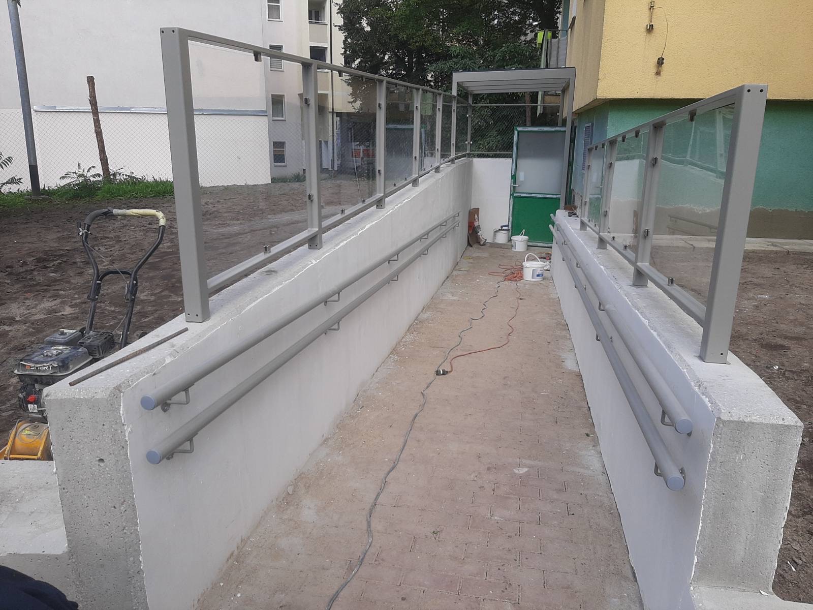 Zewnętrzna rampa dla niepełnosprawnych z szarą metalową konstrukcją i szklanymi panelami ochronnymi oraz podwójną poręczą, widoczny budynek w tle i sprzęt budowlany.